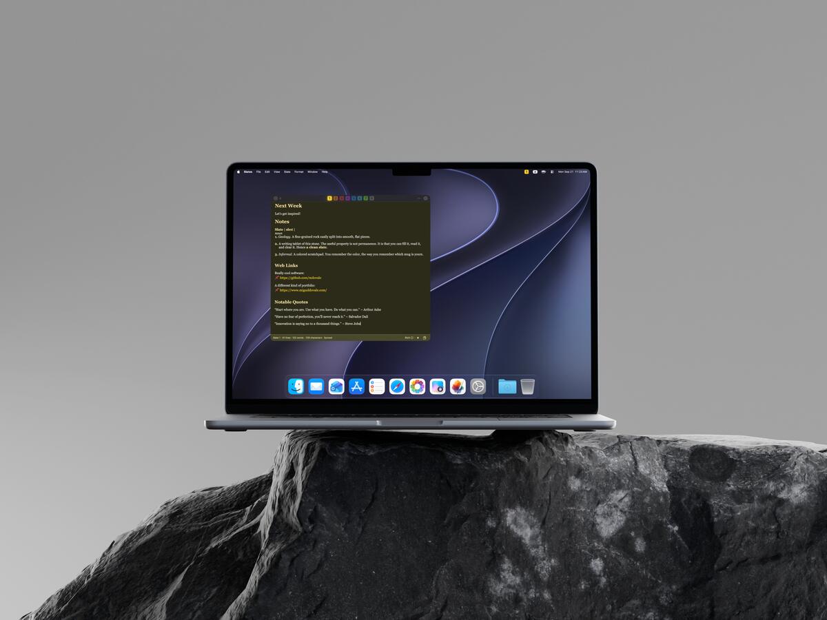 A MacBook Air balanced on a dark natural rock against a gray backdrop, one yellow slate in focus.